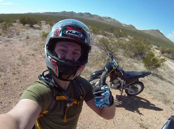 Photograph of Colton giving thumbs up while standing in the desert with a motorcycle in the background